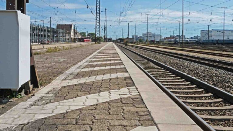 Em um dia claro, uma plataforma de estação de trem vazia aguarda, exibindo vários trilhos ferroviários. Linhas de energia e uma caixa de sinalização se destacam em primeiro plano, sutilmente reminiscentes de como os produtos da China navegam em sua jornada legalmente intrincada para novos destinos.