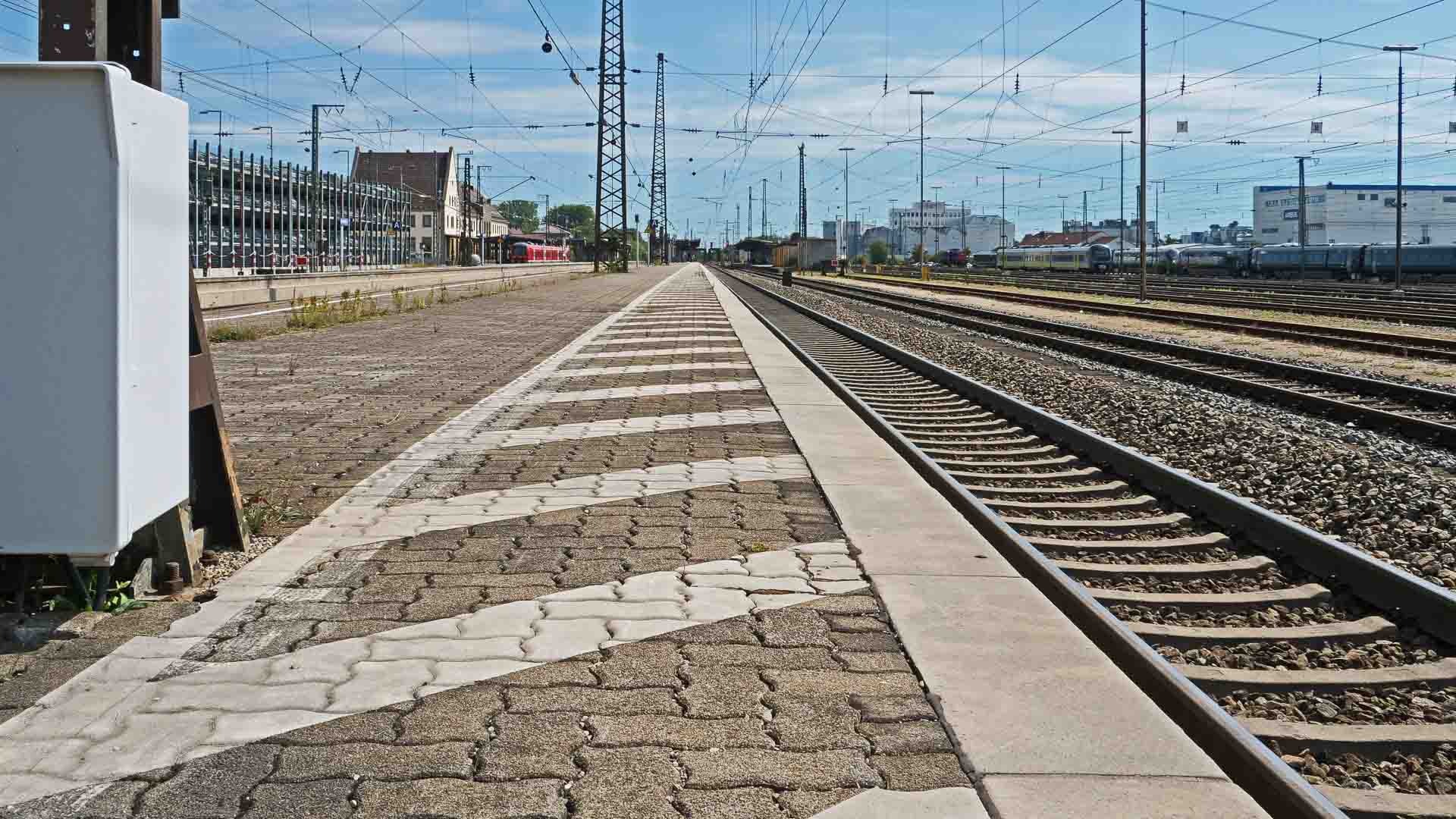 Em um dia claro, uma plataforma de estação de trem vazia aguarda, exibindo vários trilhos ferroviários. Linhas de energia e uma caixa de sinalização se destacam em primeiro plano, sutilmente reminiscentes de como os produtos da China navegam em sua jornada legalmente intrincada para novos destinos.