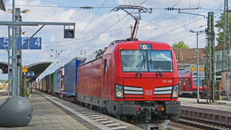 Um trem de carga vermelho com uma série de vagões de contêineres parou na plataforma de uma estação de trem sob um céu nublado, sugerindo o transporte de "Produtos Importados" para aqueles que estão aprendendo "Como Vender". Linhas de energia e placas de estação são visíveis, emoldurando esta cena de comércio internacional.