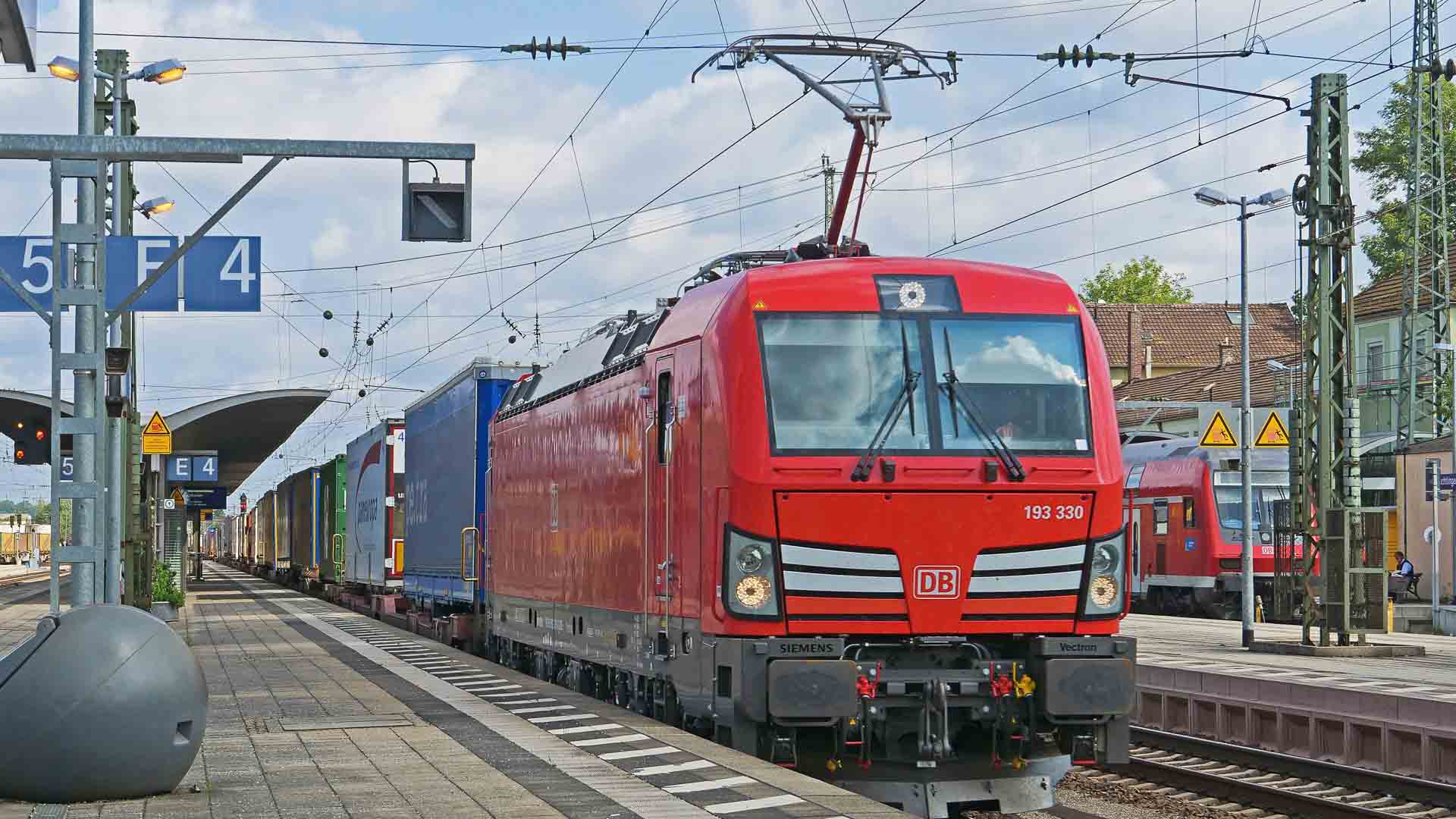 Um trem de carga vermelho com uma série de vagões de contêineres parou na plataforma de uma estação de trem sob um céu nublado, sugerindo o transporte de "Produtos Importados" para aqueles que estão aprendendo "Como Vender". Linhas de energia e placas de estação são visíveis, emoldurando esta cena de comércio internacional.