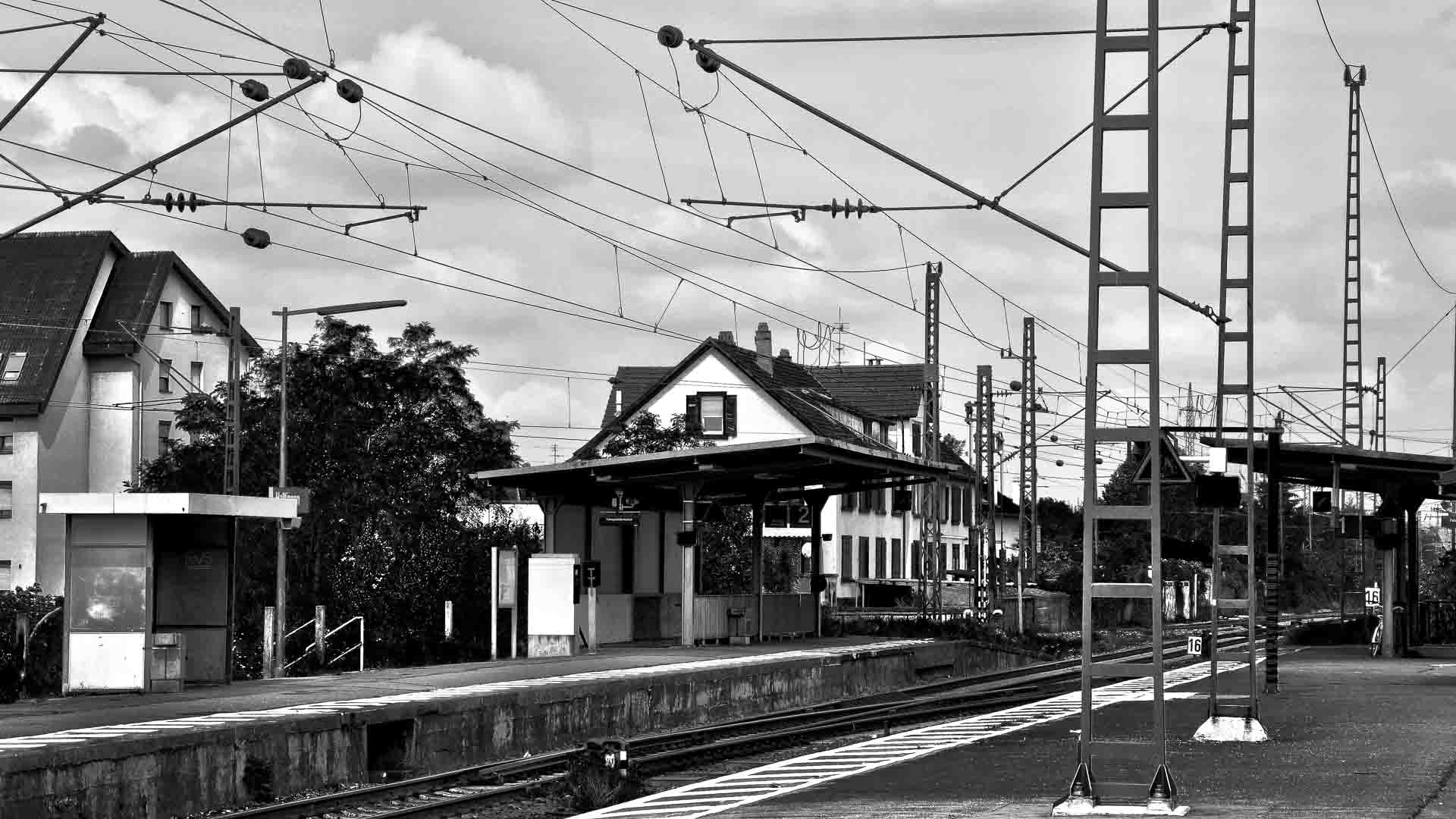 Uma imagem em preto e branco de uma plataforma de estação de trem com fios aéreos e alguns prédios ao fundo, lembrando os centros movimentados onde as mercadorias eram tradicionalmente trocadas antes de práticas modernas como importar da China alterarem as cadeias de suprimentos. Nenhum trem está presente.