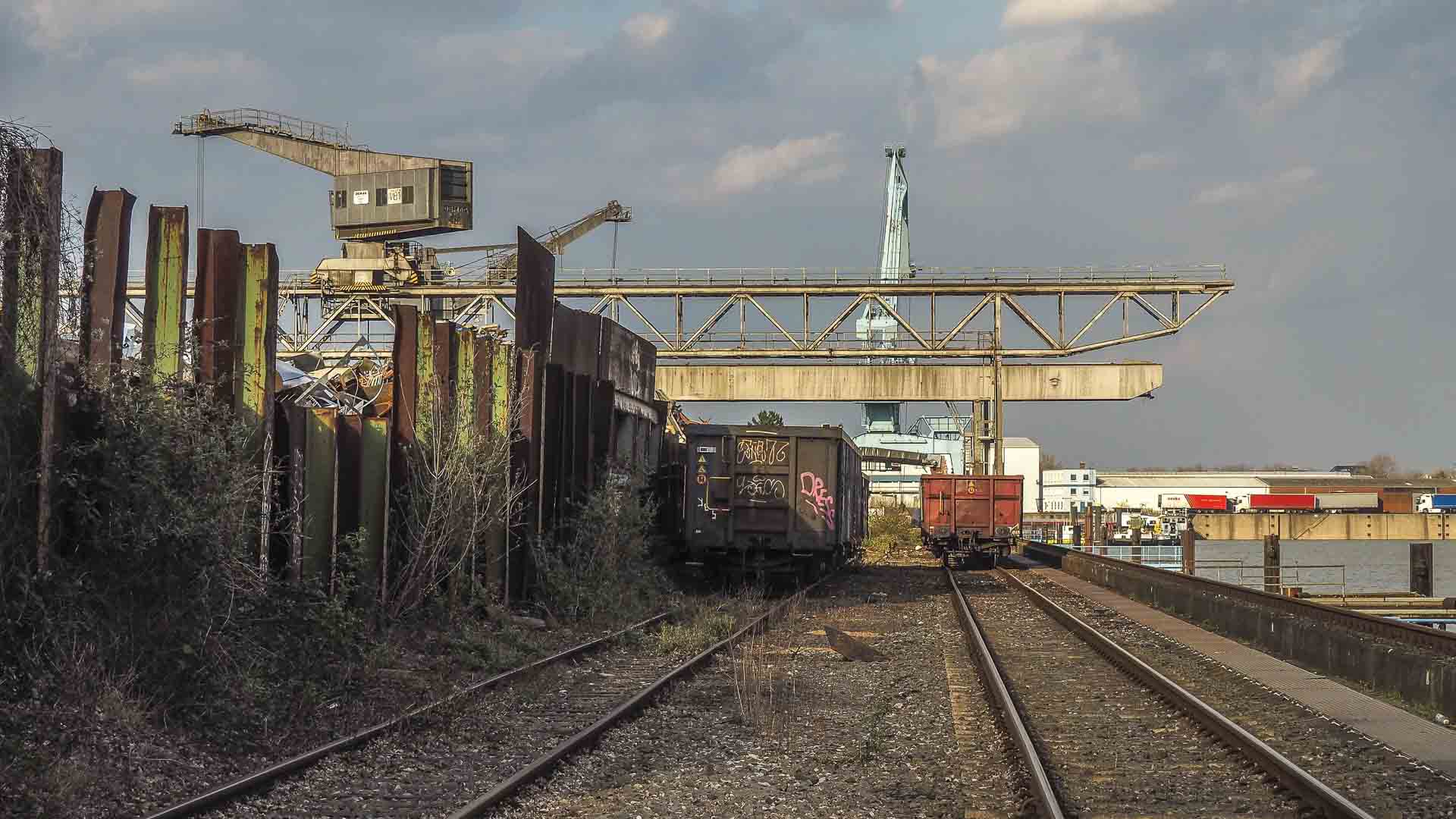 Trilhos ferroviários industriais abandonados com vagões de carga e estruturas enferrujadas, incluindo guindastes, sussurram histórias de comércio passado contra um céu nublado, como se esperassem para calcular processos de importação mais uma vez.