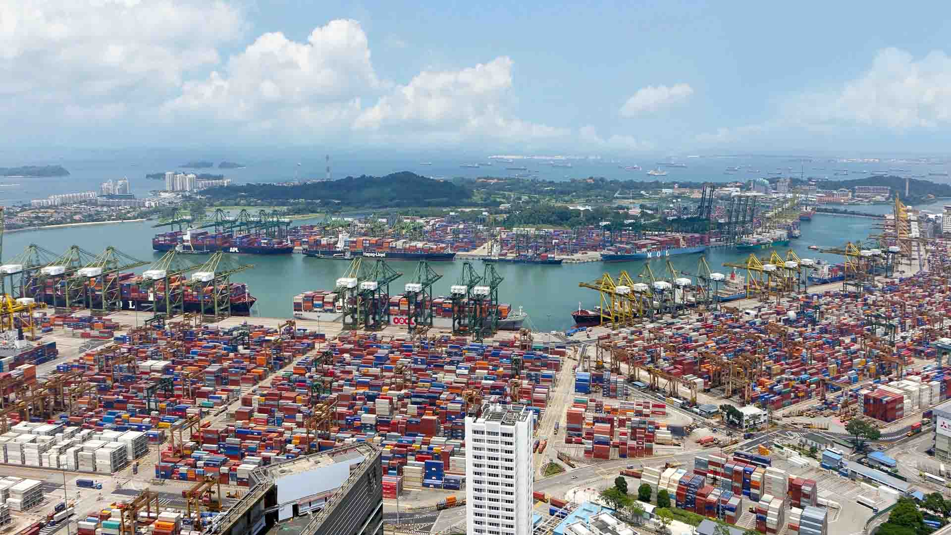 Vista aérea de um porto movimentado, ilustrando o processo passo a passo do desembaraço aduaneiro, com vários contêineres, guindastes e navios atracados ao longo do canal, tendo como pano de fundo o céu azul e ilhas distantes.