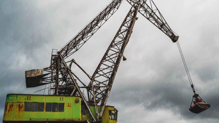 Um grande guindaste industrial com uma cabine verde enferrujada ergue uma garra de metal contra um céu nublado, simbolizando o papel vital que essas máquinas desempenham na importação e no suporte de pequenos negócios.