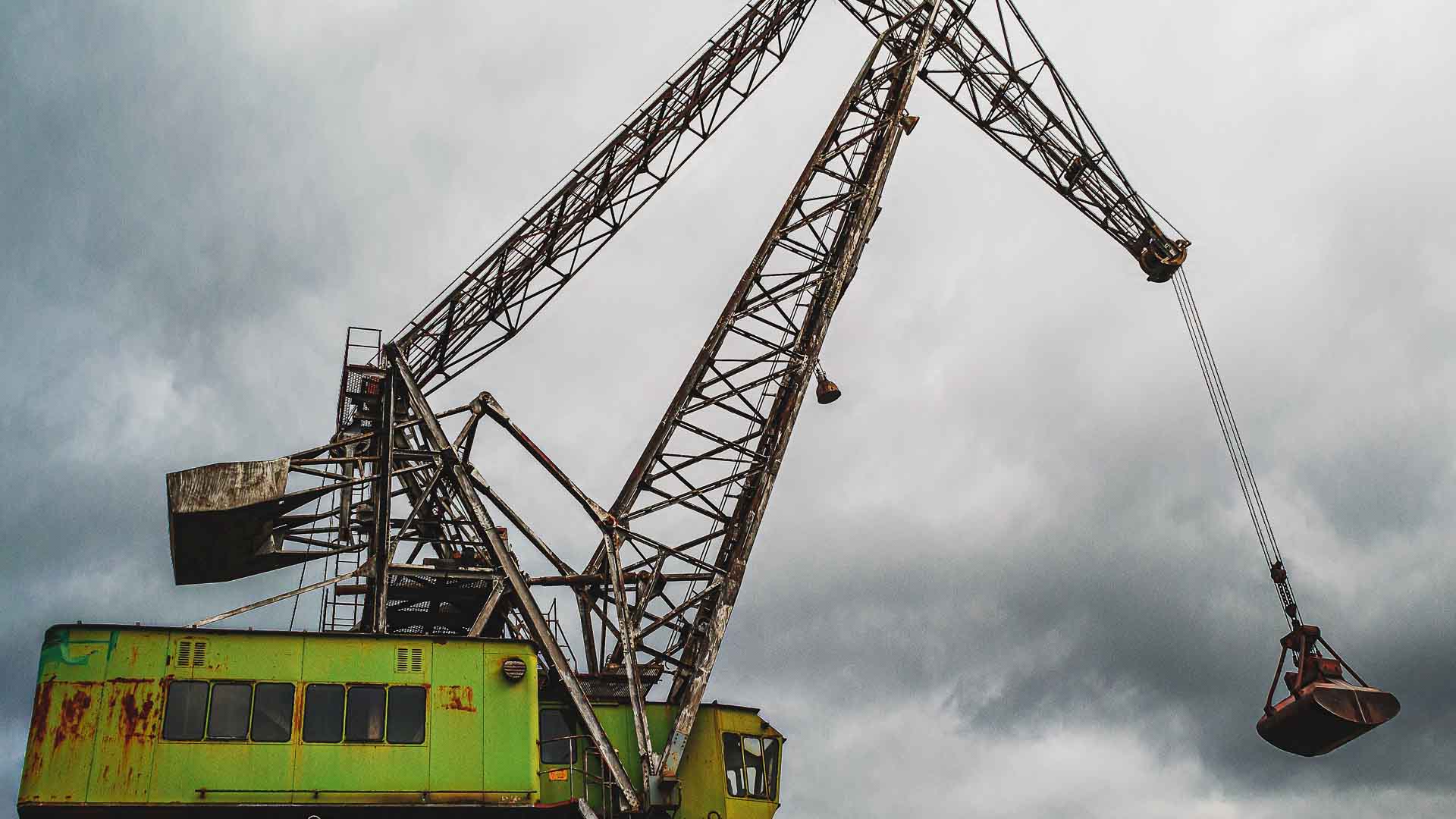 Um grande guindaste industrial com uma cabine verde enferrujada ergue uma garra de metal contra um céu nublado, simbolizando o papel vital que essas máquinas desempenham na importação e no suporte de pequenos negócios.