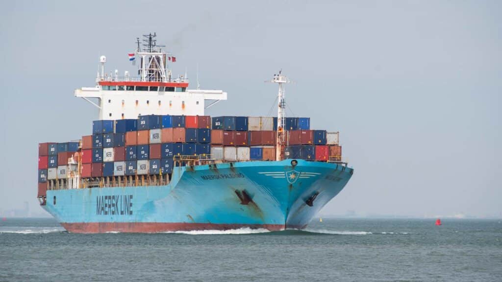 Um grande navio de carga carregado com contêineres coloridos empilhados navega por um corpo de água sob um céu limpo, lembrando uma viagem à China.
