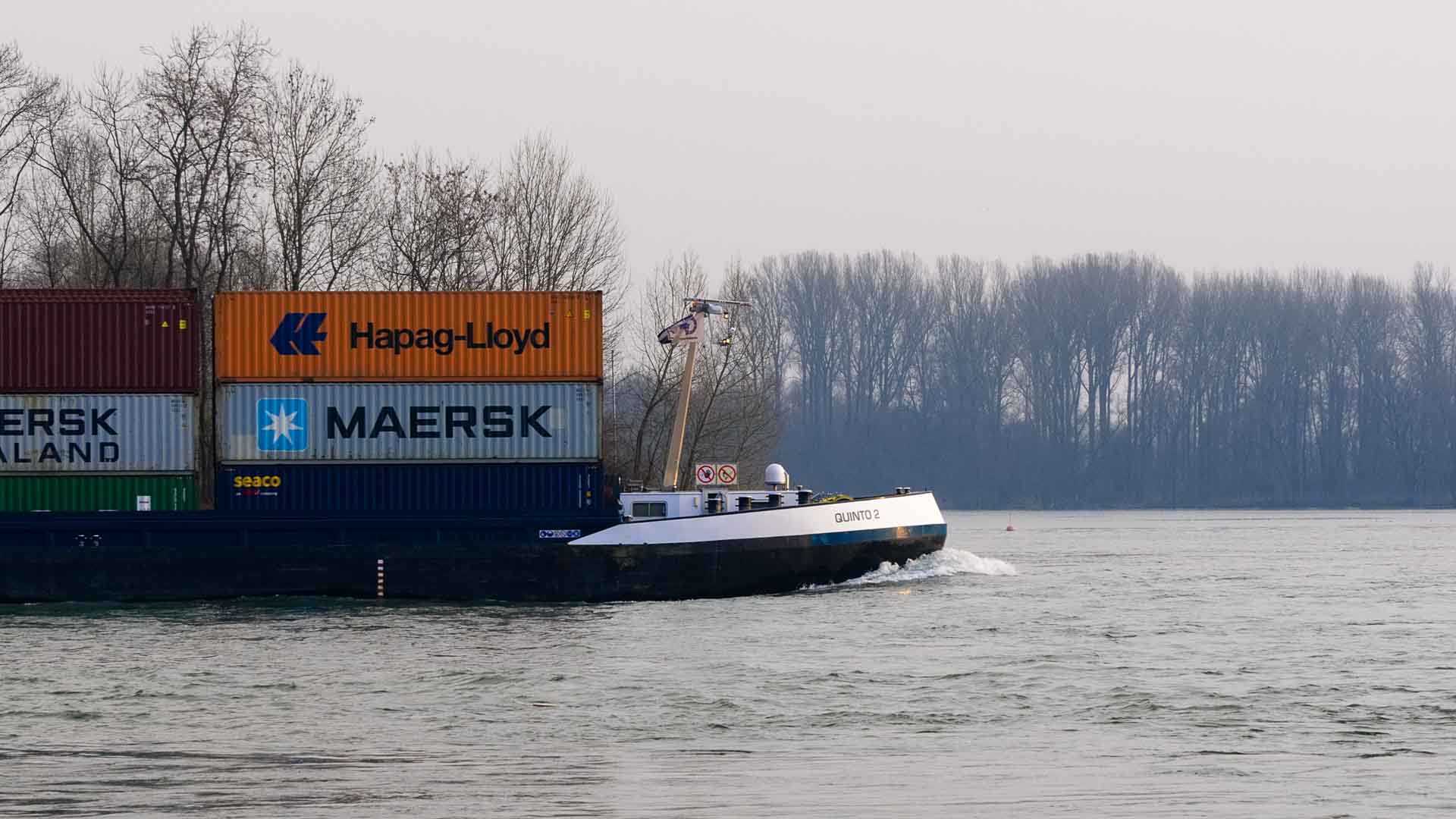 Um navio de carga transporta contêineres da Maersk e da Hapag-Lloyd ao longo de um rio, navegando com a precisão de um prático, cercado por árvores nuas sob um céu cinza durante sua longa viagem à China.