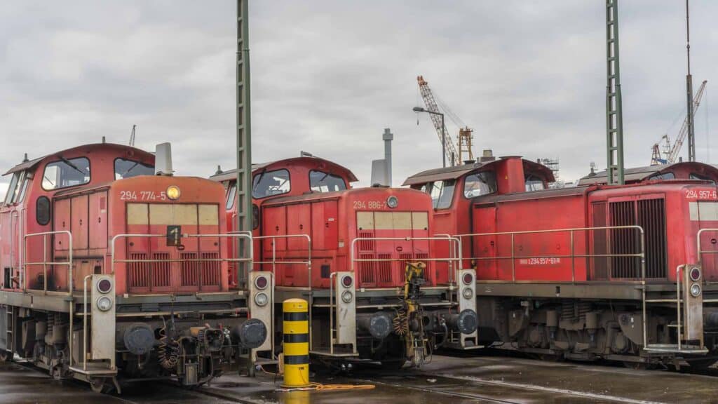 Três locomotivas a diesel vermelhas estão estacionadas em um pátio ferroviário, onde estruturas industriais e guindastes se erguem sob um céu nublado, representando a segurança vital para uma operação eficiente.