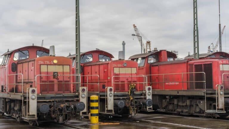 Três locomotivas a diesel vermelhas estão estacionadas em um pátio ferroviário, onde estruturas industriais e guindastes se erguem sob um céu nublado, representando a segurança vital para uma operação eficiente.