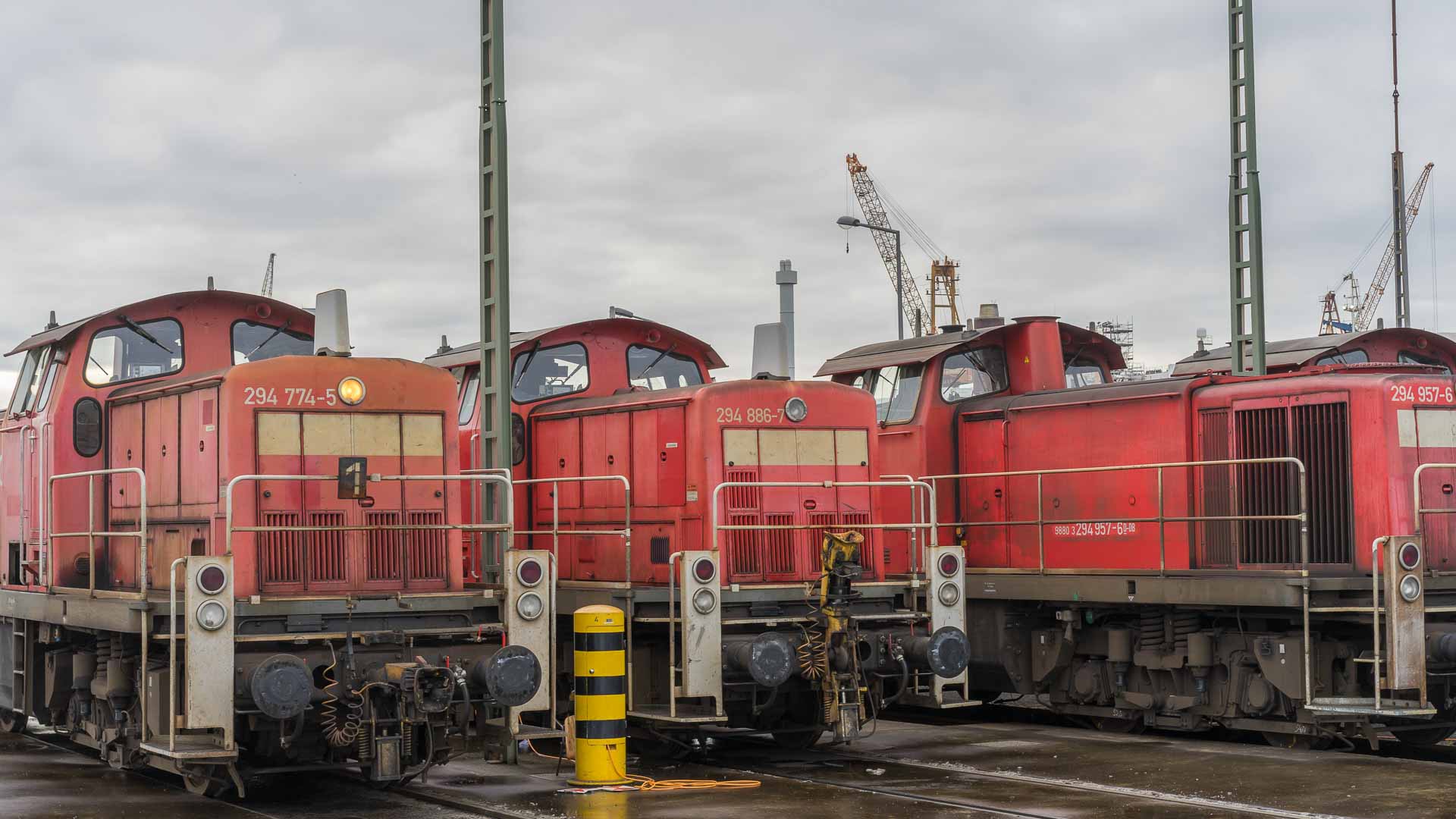 Três locomotivas a diesel vermelhas estão estacionadas em um pátio ferroviário, onde estruturas industriais e guindastes se erguem sob um céu nublado, representando a segurança vital para uma operação eficiente.