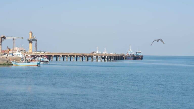 Uma cena tranquila de porto marítimo apresenta um longo píer se estendendo em direção ao calmo mar azul, lembrando os pitorescos portos da China. Barcos e navios estão atracados pacificamente enquanto um pássaro solitário navega graciosamente pelo céu, convidando a saborear esse momento de beleza serena.