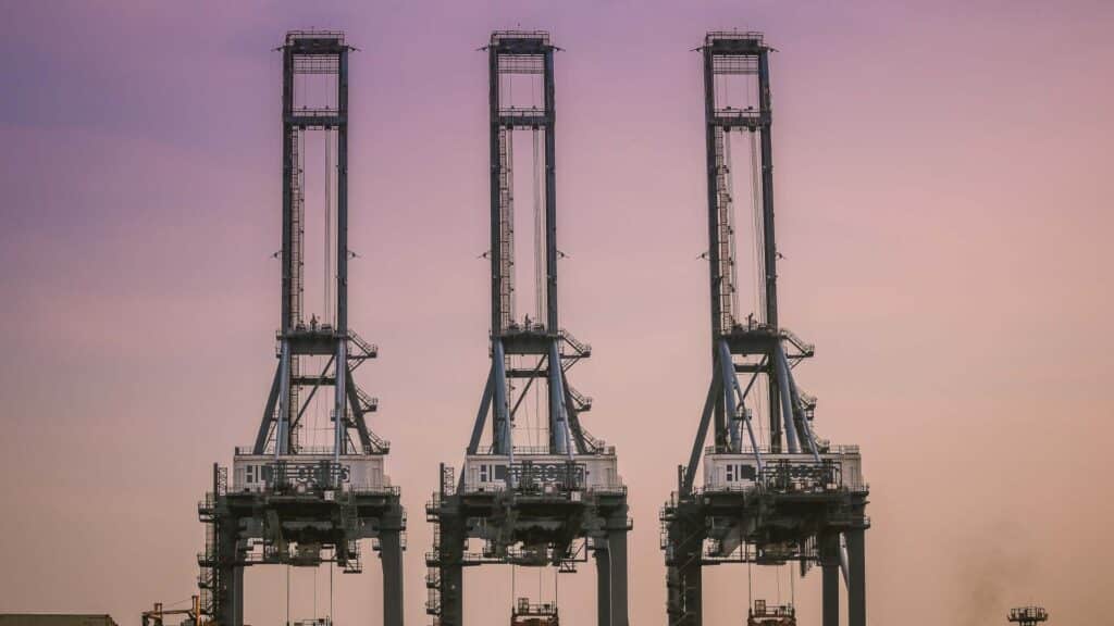 Três grandes guindastes industriais estão alinhados contra um céu noturno roxo e laranja, orquestrando com eficiência a importação de mercadorias da China no porto, garantindo operações tranquilas e maximizando o lucro para todos os envolvidos.
