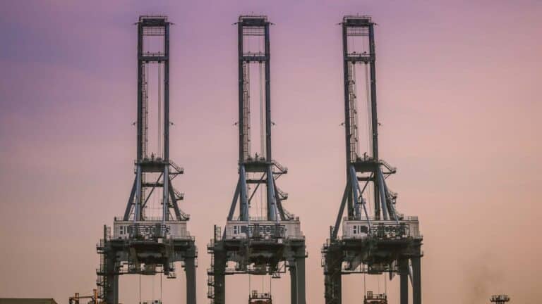 Três grandes guindastes industriais estão alinhados contra um céu noturno roxo e laranja, orquestrando com eficiência a importação de mercadorias da China no porto, garantindo operações tranquilas e maximizando o lucro para todos os envolvidos.