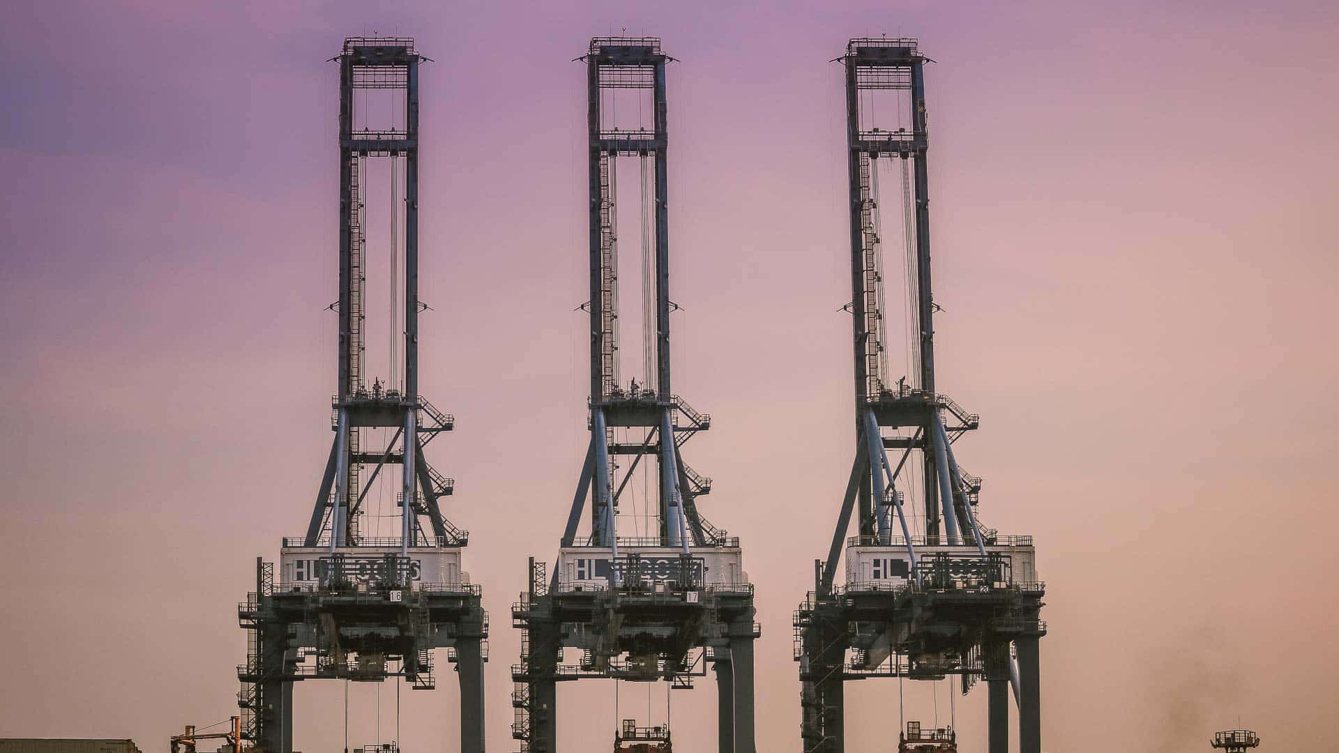 Três grandes guindastes industriais estão alinhados contra um céu noturno roxo e laranja, orquestrando com eficiência a importação de mercadorias da China no porto, garantindo operações tranquilas e maximizando o lucro para todos os envolvidos.