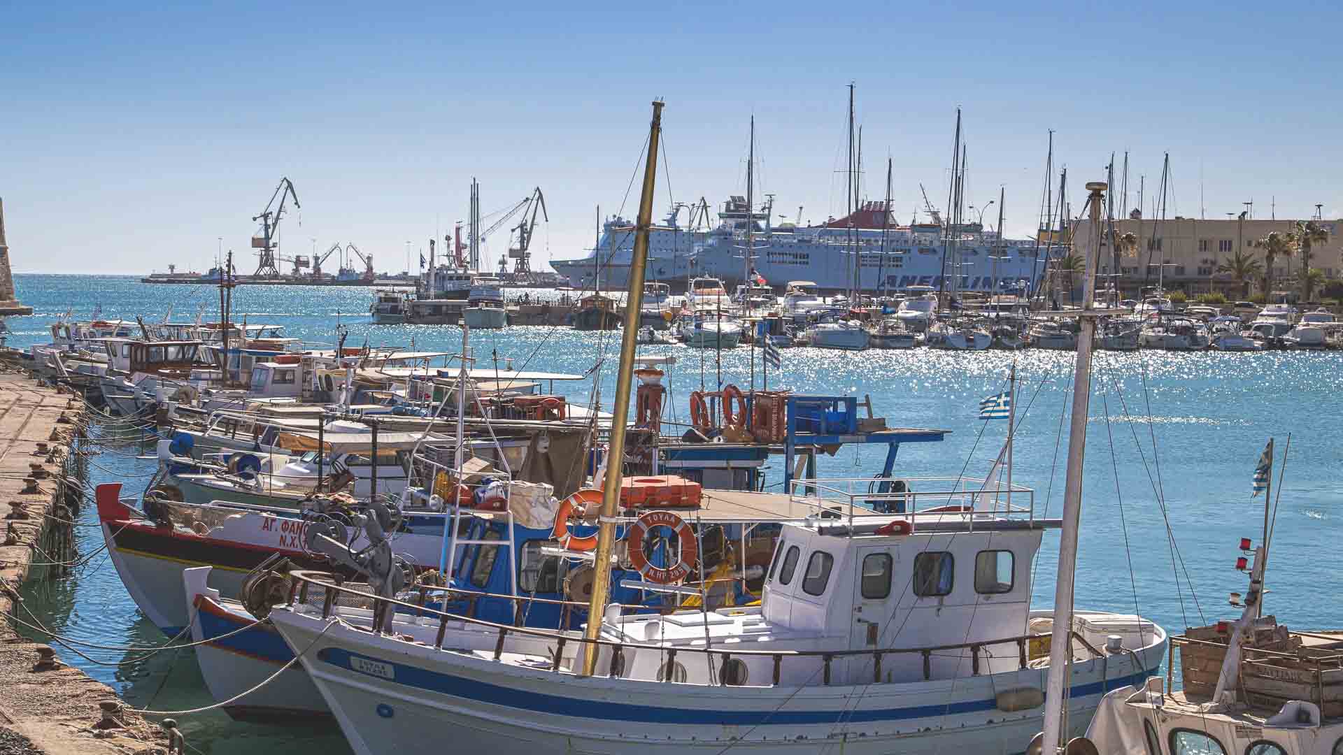 Um porto movimentado com vários barcos atracados ao longo do píer e grandes navios, prontos para embarcação, surgindo ao fundo sob um céu azul claro.