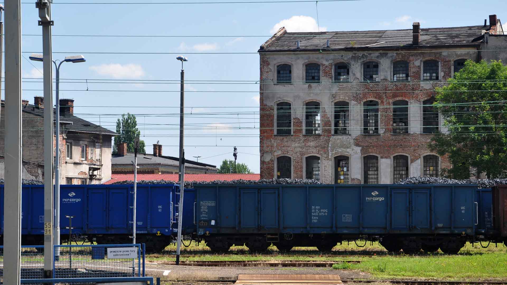 Vagões de trem de carga azuis carregados com carvão estão em um pátio ferroviário ao lado de um antigo prédio de tijolos com janelas em arco. Contra o pano de fundo de um céu azul claro e linhas de energia estendidas, a cena sugere rotas comerciais movimentadas que também podem facilitar a importação de produtos da China.