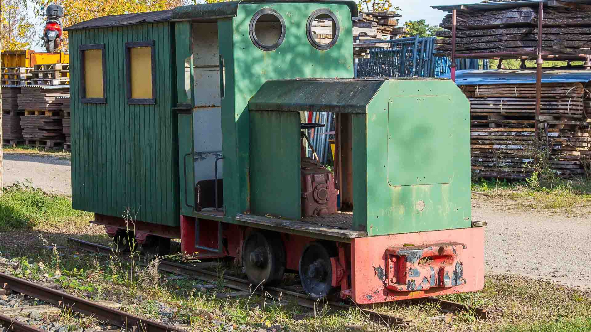 Uma pequena locomotiva industrial verde e vermelha com duas janelas redondas está estacionada em um trilho no local de Cresceram, cercada por pilhas de materiais.