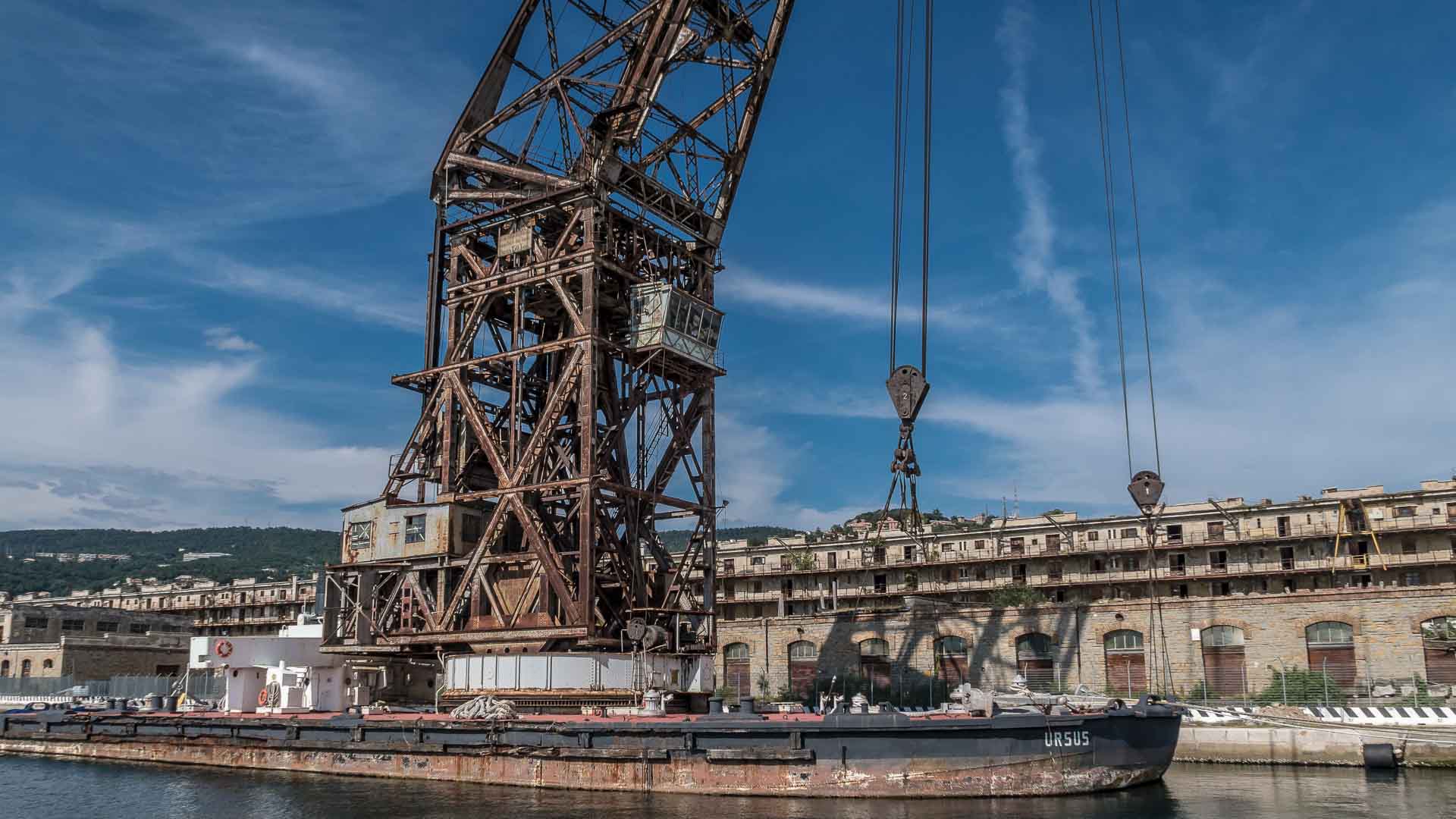 Um antigo guindaste industrial em uma plataforma flutuante em uma doca, contra edifícios e colinas sob um céu azul claro, permanece como uma testemunha silenciosa da movimentada atividade de importação que prospera aqui.