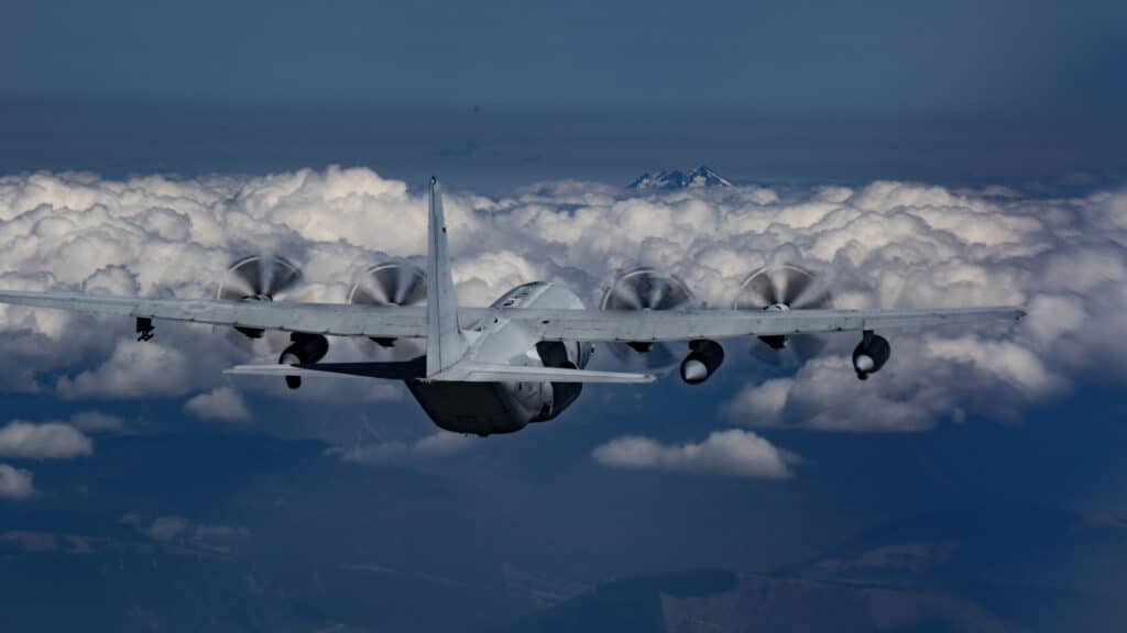 Uma aeronave militar com quatro hélices voa acima de uma densa camada de nuvens, empregando estratégias para navegar pelos céus, com um pico de montanha distante visível ao fundo.