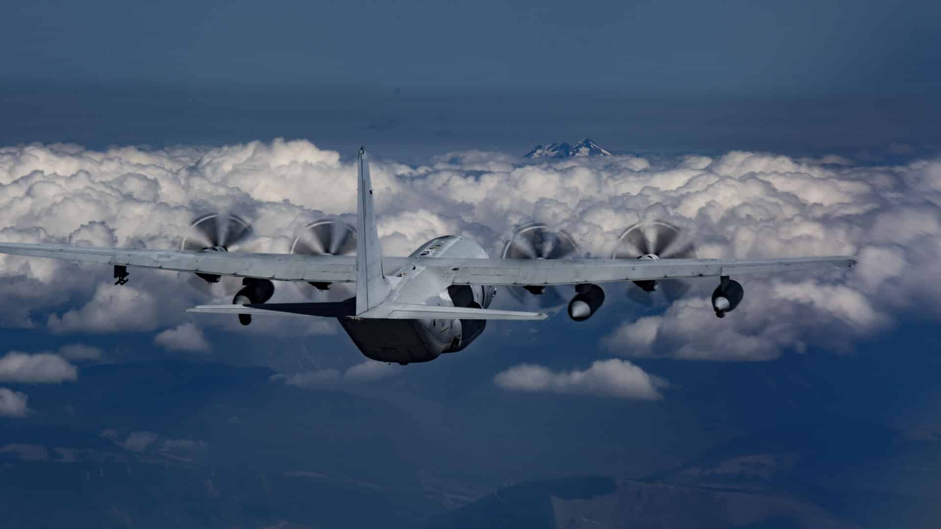 Uma aeronave militar com quatro hélices voa acima de uma densa camada de nuvens, empregando estratégias para navegar pelos céus, com um pico de montanha distante visível ao fundo.