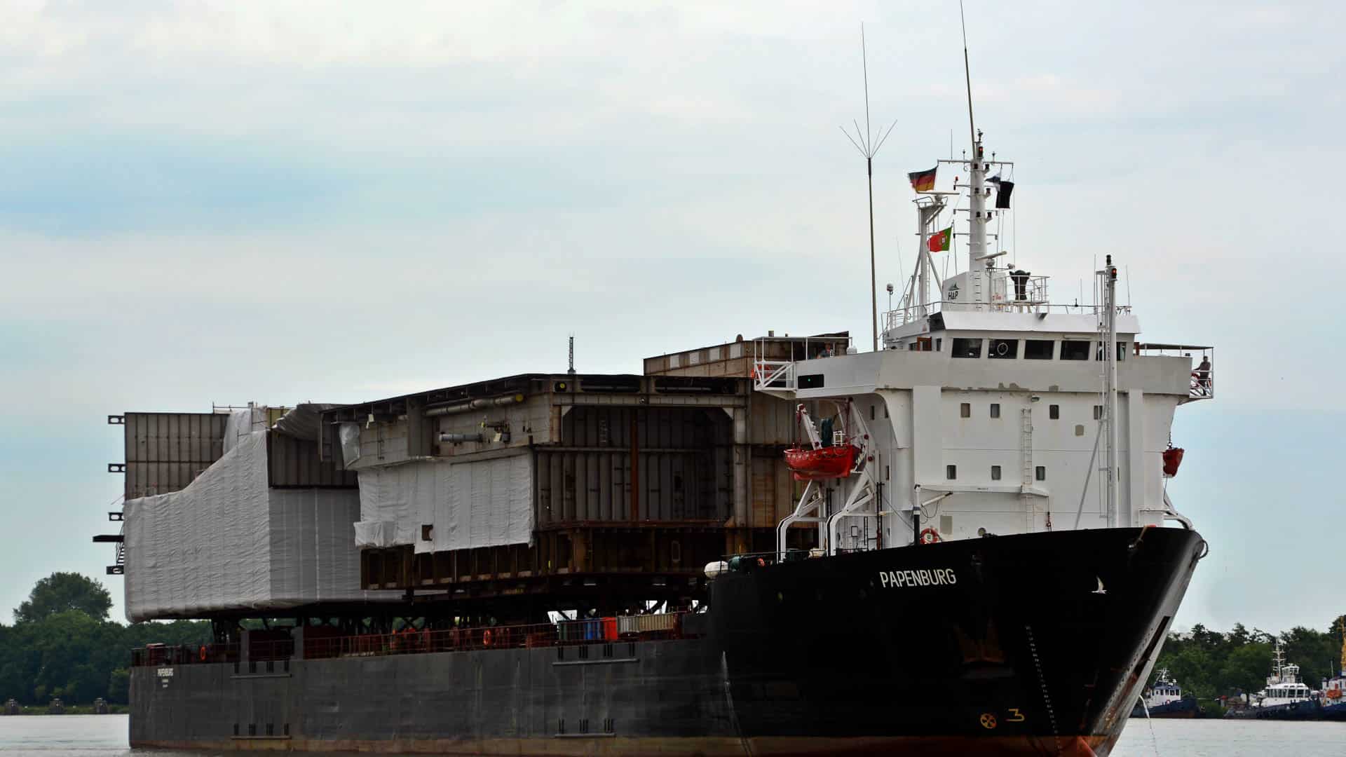 Um navio de carga, carregado com grandes equipamentos industriais, navega pelo rio sob um céu nublado, enquanto árvores se alinham ao fundo. O complexo processo de "desembaraço aduaneiro" é essencial para garantir uma passagem suave, assim como a natureza encena seu cenário sereno.