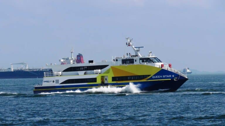 Uma balsa amarela e azul chamada "Queen Star 2" desliza pela água, simbolizando o sucesso em viagens marítimas, com navios de carga visíveis ao fundo sob um céu parcialmente nublado.