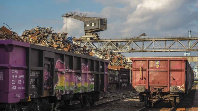 Dois vagões de trem de carga nos trilhos ao lado de uma pilha de sucata contam uma história de expectativas industriais para 2024. Uma correia transportadora trabalha incansavelmente com uma estrutura industrial ao fundo sob um céu nublado, sugerindo o potencial aumento de indústrias que se avizinha no horizonte.