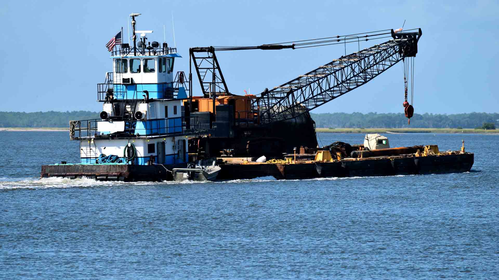 Uma barcaça fluvial com guindaste, perfeita para tarefas de importação, navega por águas calmas sob um céu azul claro.