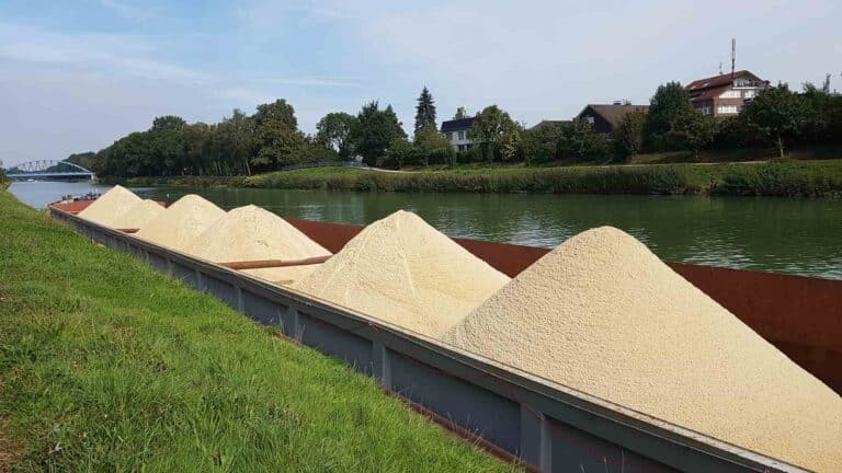 Uma barcaça em um canal carrega vários montes de um material granular de cor clara, como os bens inestimáveis transportados pela rede do Alibaba. Casas e árvores se alinham na margem oposta, criando uma cena pacífica.