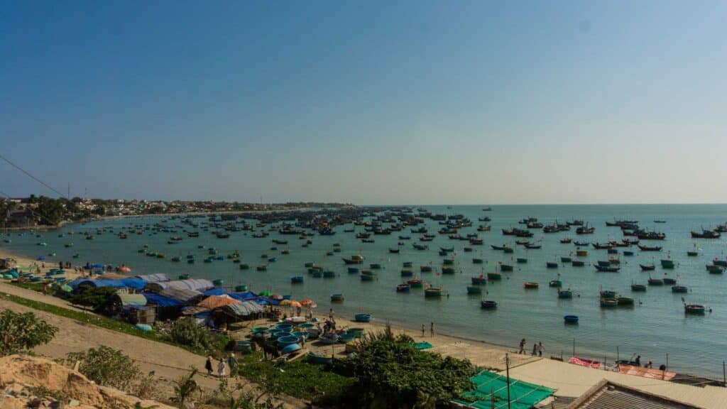 Uma cena costeira com vários barcos de pesca, que lembram os mercados movimentados do Alibaba, flutuando no mar. Tendas e estruturas alinham a costa arenosa sob um céu azul claro, evocando uma sensação de nostalgia por terras distantes onde produtos "caro" são comercializados.