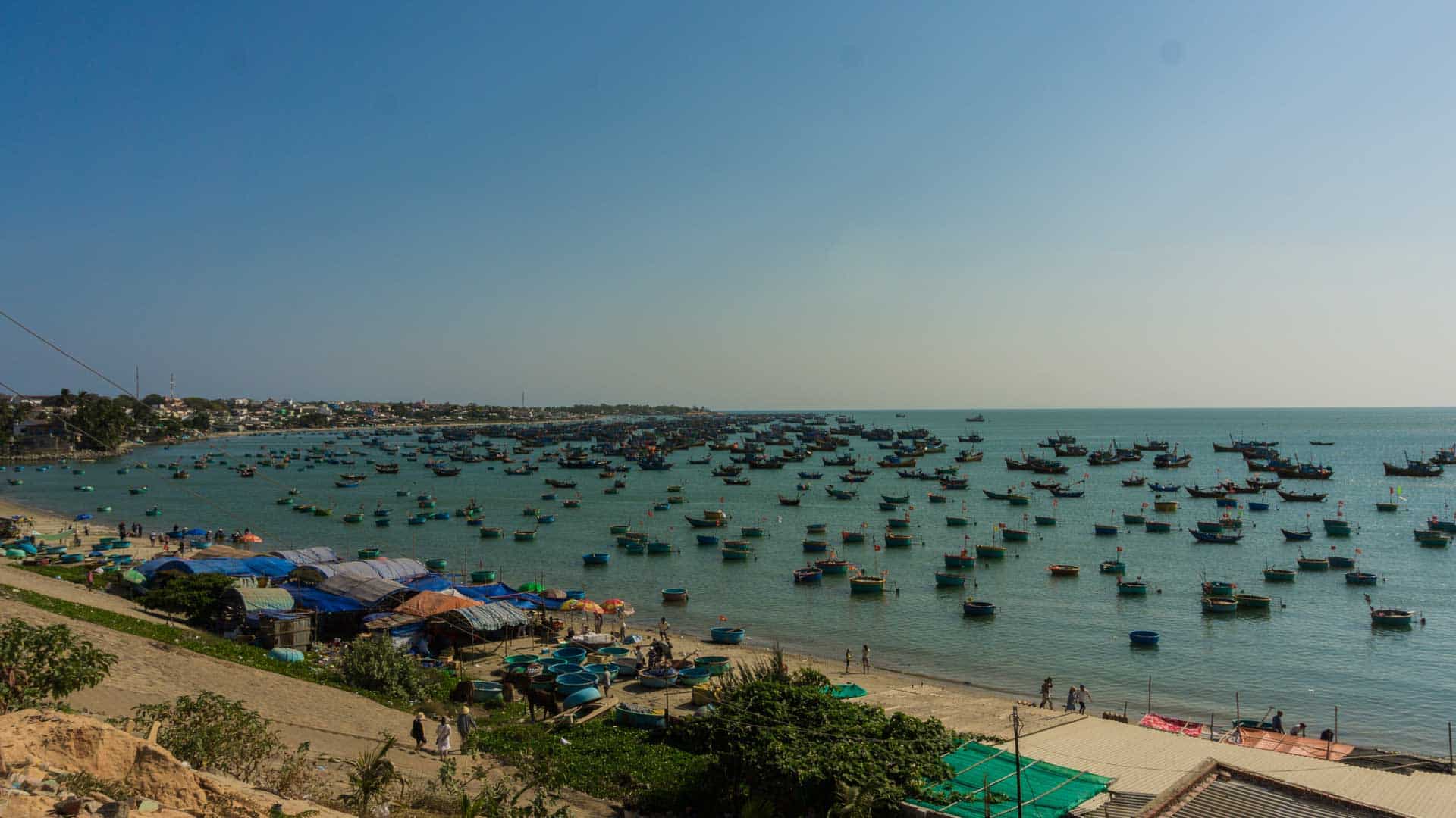 Uma cena costeira com vários barcos de pesca, que lembram os mercados movimentados do Alibaba, flutuando no mar. Tendas e estruturas alinham a costa arenosa sob um céu azul claro, evocando uma sensação de nostalgia por terras distantes onde produtos "caro" são comercializados.