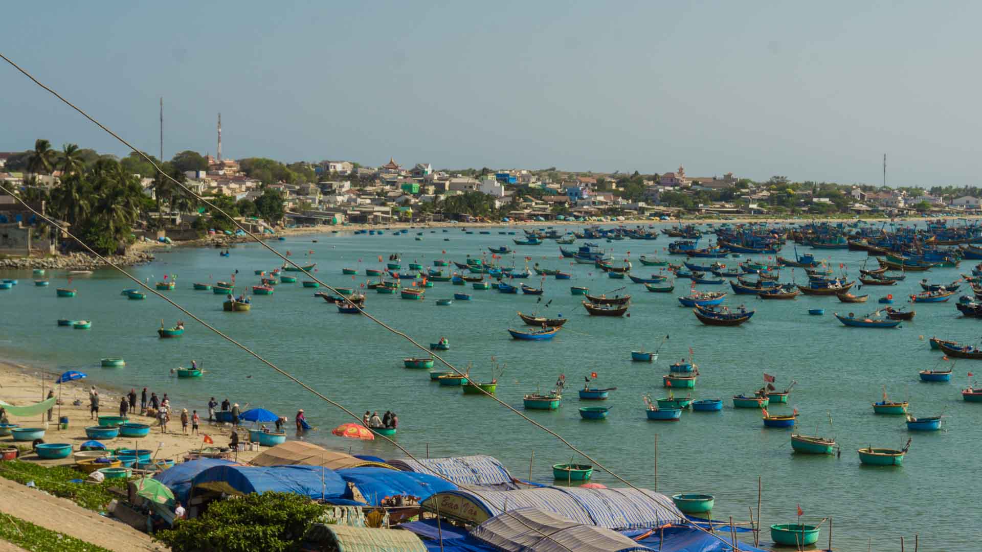 Uma cidade costeira movimentada com pequenos barcos e tendas alinhadas na costa, emoldurada por colinas e prédios à distância sob um céu limpo. Perfeito para iniciantes que buscam explorar a vida marítima conforme nos aproximamos de 2025.