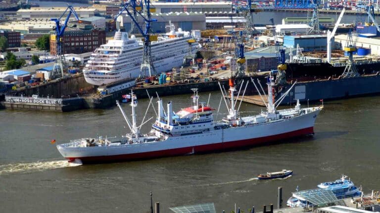 Um navio de carga branco e vermelho, transportando mercadorias vitais para importadores, navega por um porto movimentado. Ele é cercado por guindastes, armazéns, um navio de cruzeiro e barcos menores — todos movimentados como capítulos de um guia de certificação.