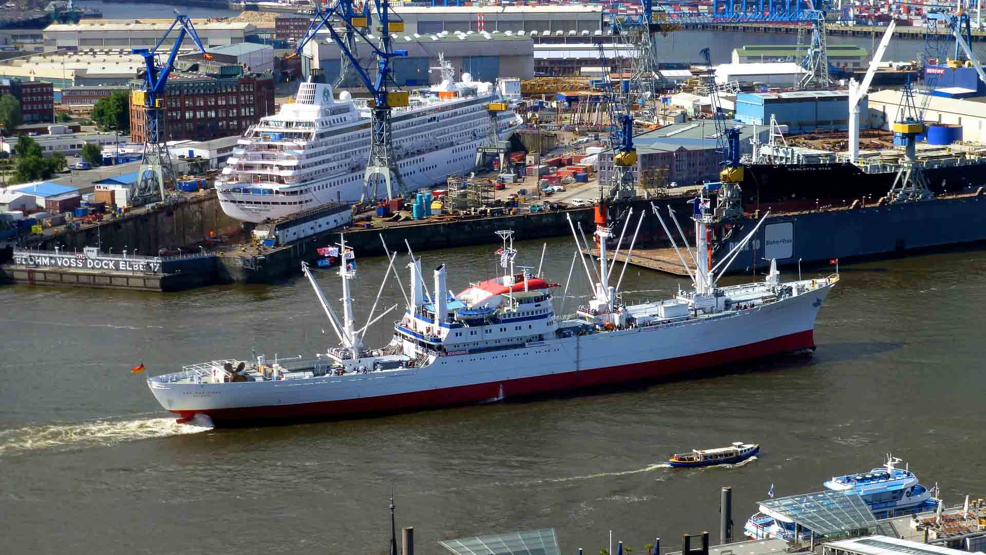 Um navio de carga branco e vermelho, transportando mercadorias vitais para importadores, navega por um porto movimentado. Ele é cercado por guindastes, armazéns, um navio de cruzeiro e barcos menores — todos movimentados como capítulos de um guia de certificação.