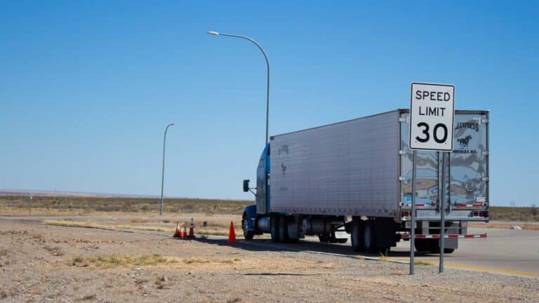 Um caminhão grande dirige em uma estrada em uma paisagem desértica, talvez transportando mercadorias da China. Uma placa de limite de velocidade exibe 30 mph, e cones de trânsito são visíveis perto do caminhão.