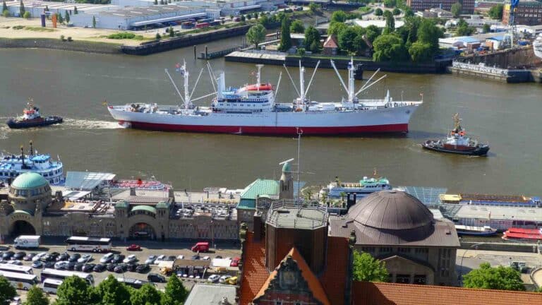 Vista aérea de um grande navio cargueiro branco e vermelho navegando no rio, cercado por rebocadores. Esta cena movimentada simboliza a importação em expansão em 2025, com uma área urbana apresentando vários edifícios e vegetação ao fundo, sugerindo novos negócios no horizonte.