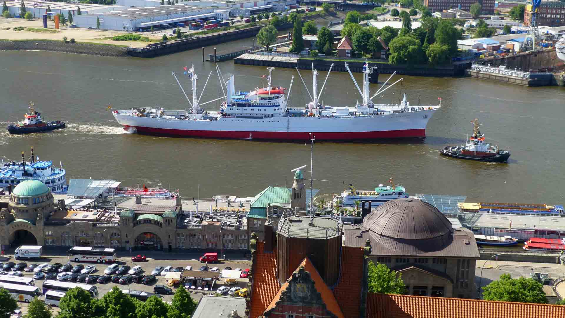 Vista aérea de um grande navio cargueiro branco e vermelho navegando no rio, cercado por rebocadores. Esta cena movimentada simboliza a importação em expansão em 2025, com uma área urbana apresentando vários edifícios e vegetação ao fundo, sugerindo novos negócios no horizonte.