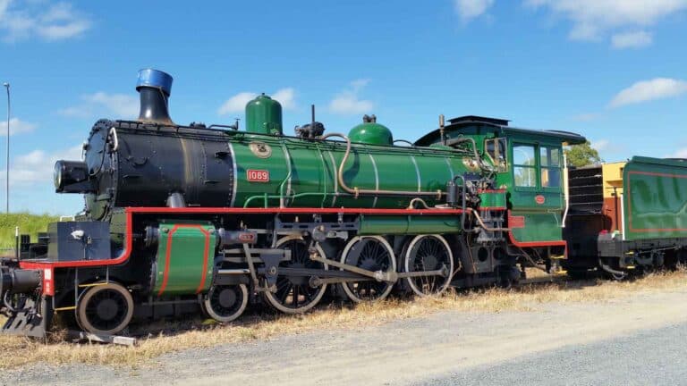 Uma locomotiva a vapor verde com detalhes em vermelho e preto está estacionada em um caminho de cascalho sob um céu azul, oferecendo uma viagem atemporal que lembra a rica história da importação.