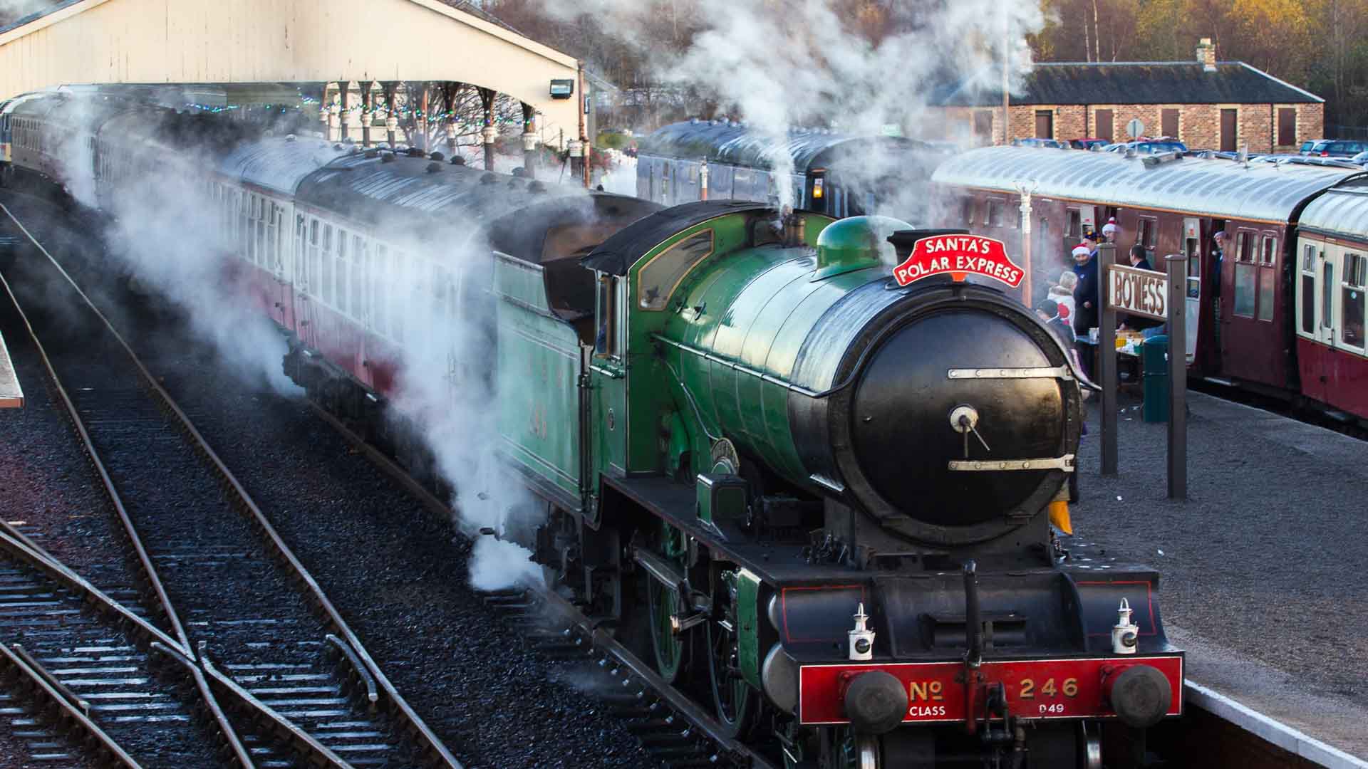 Uma locomotiva a vapor com o nome "Santa's Polar Express" está majestosamente na estação, liberando uma nuvem de vapor enquanto carruagens de passageiros vintage aguardam. As pessoas se reúnem nas proximidades, ansiosas para embarcar em sua aventura de férias importada.