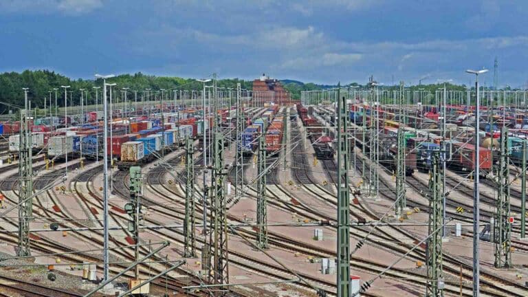 Um grande pátio ferroviário com vários trilhos está cheio de contêineres empilhados aguardando produtos importantes, enquanto vagões ficam sob um céu nublado, garantindo que tudo atenda à certificação necessária.