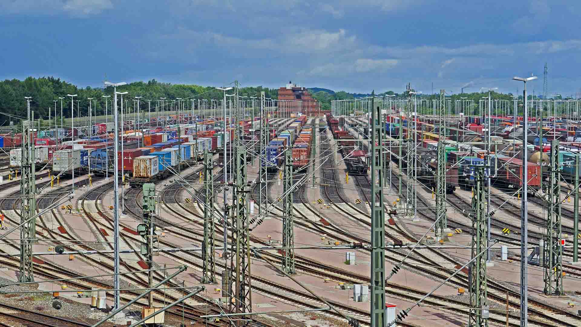 Um grande pátio ferroviário com vários trilhos está cheio de contêineres empilhados aguardando produtos importantes, enquanto vagões ficam sob um céu nublado, garantindo que tudo atenda à certificação necessária.