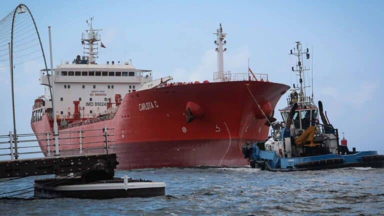 Um grande navio cargueiro vermelho, símbolo de importação, está sendo guiado por um rebocador menor por uma hidrovia. Uma ponte é parcialmente visível no lado esquerdo da imagem, sugerindo o impacto da tecnologia na navegação marítima até 2025.