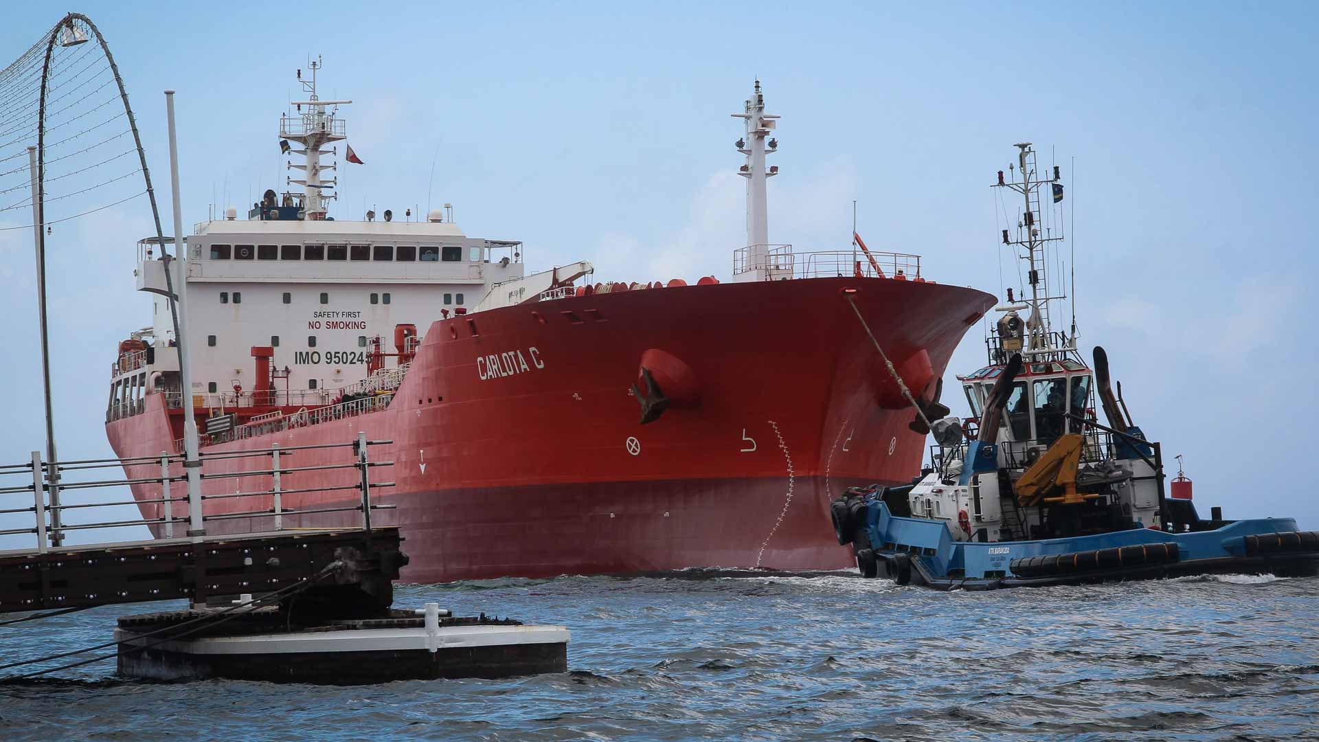 Um grande navio cargueiro vermelho, símbolo de importação, está sendo guiado por um rebocador menor por uma hidrovia. Uma ponte é parcialmente visível no lado esquerdo da imagem, sugerindo o impacto da tecnologia na navegação marítima até 2025.