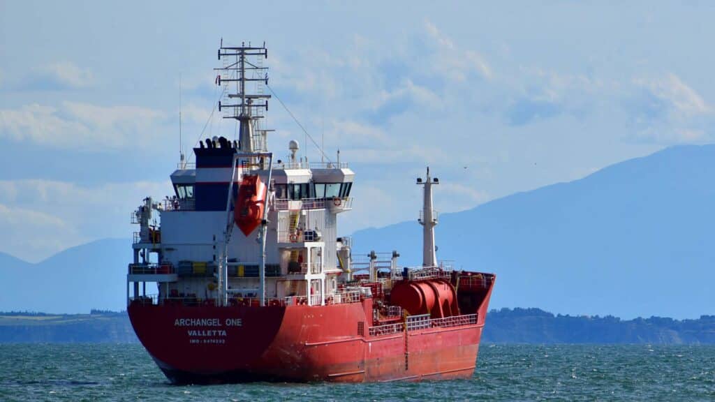 O navio cargueiro vermelho, Archangel One, desliza serenamente nas águas cintilantes com montanhas majestosas ao fundo. Esta embarcação desempenha um papel crucial nas rotas comerciais globais entre a China e outras nações.