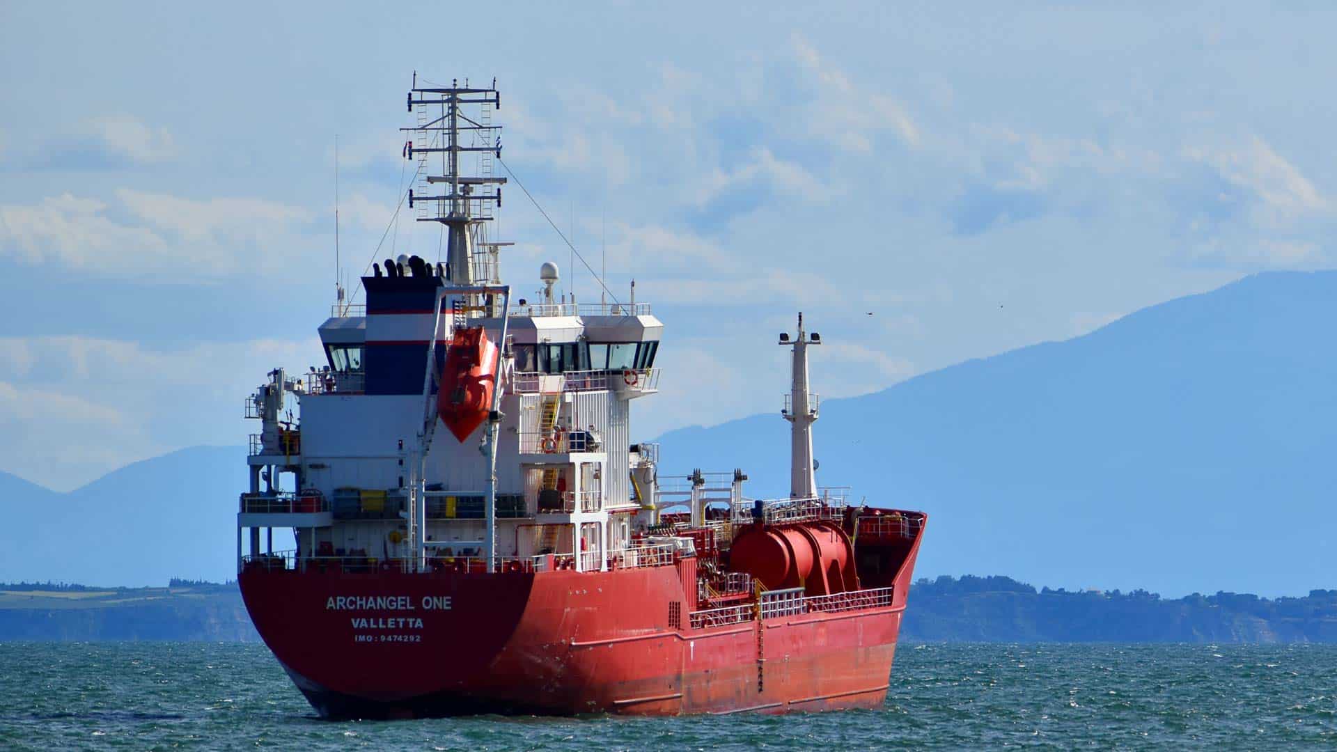 O navio cargueiro vermelho, Archangel One, desliza serenamente nas águas cintilantes com montanhas majestosas ao fundo. Esta embarcação desempenha um papel crucial nas rotas comerciais globais entre a China e outras nações.