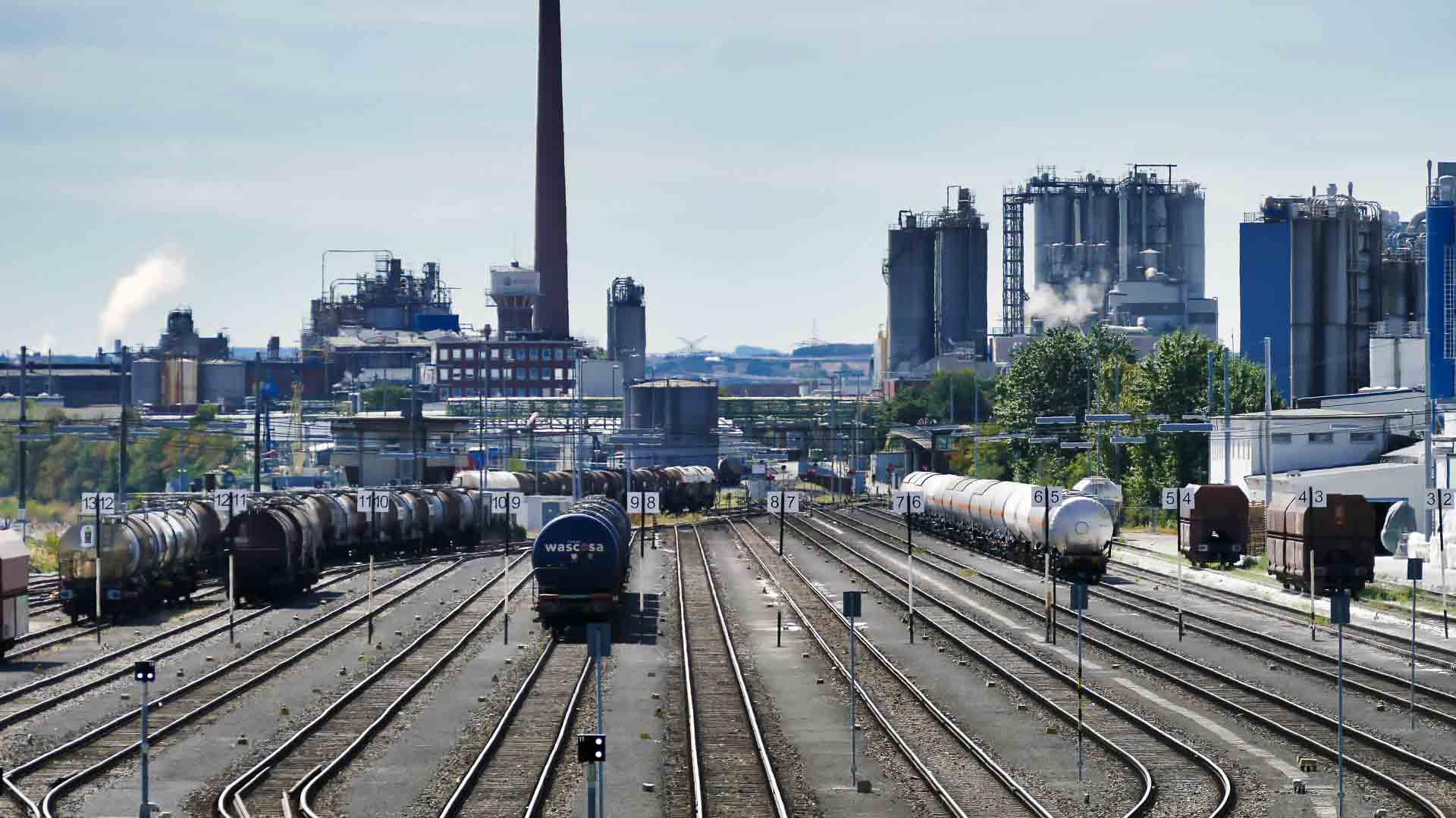 Em um pátio ferroviário industrial com vários trilhos de trem, trens de carga transportando vagões-tanque se movem com eficiência. Enquanto fábricas e chaminés se erguem ao fundo sob um céu limpo, as empresas debatem se a importação ainda vale a pena com o dólar alto impactando os custos.