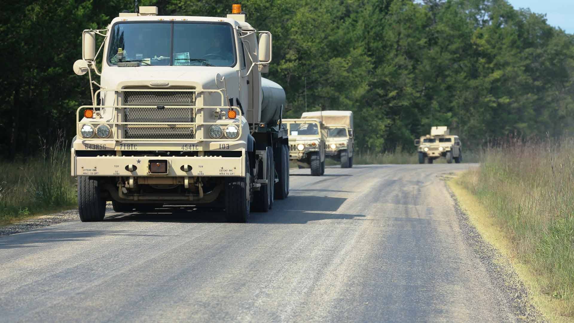 Veículos militares circulam em uma estrada rural, demonstrando precisão e força, muito parecido com os processos de importação estratégica que permitem sua implantação. Árvores se alinham em ambos os lados do caminho, como se guardassem segredos de como tais operações funcionam perfeitamente em todos os terrenos.