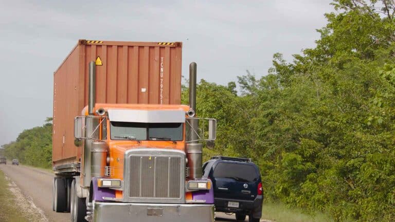 Um grande caminhão laranja com um contêiner, possivelmente para importação de ar-condicionado, trafega por uma estrada ao lado de um SUV preto e folhagem verde.