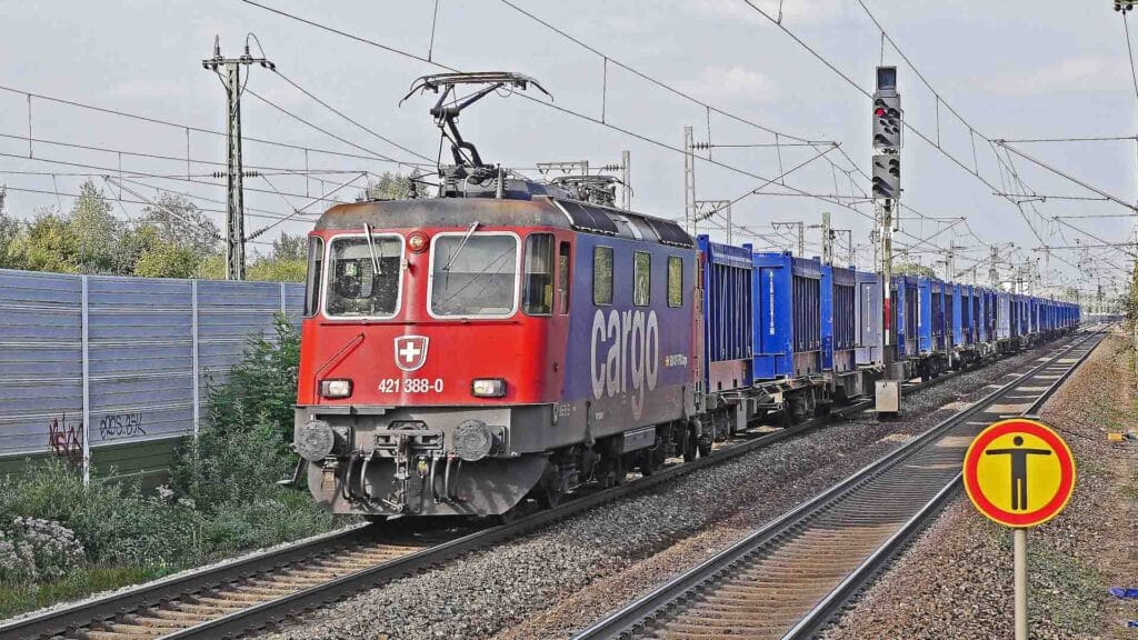 Um trem de carga suíço vermelho, possivelmente carregando itens como "Artigos de Banho", viaja em trilhos ferroviários com uma longa fila de contêineres de carga azuis. Uma placa ferroviária amarela e vermelha é visível no primeiro plano.