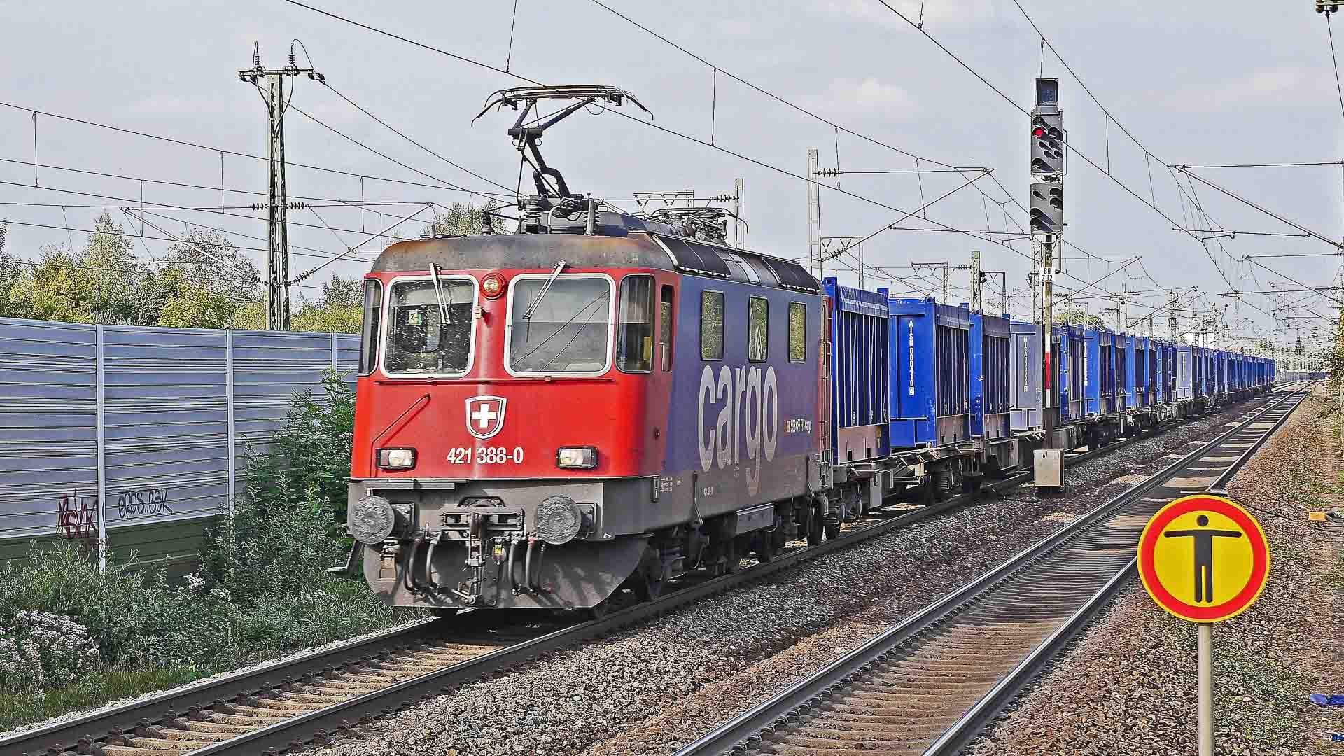 Um trem de carga suíço vermelho, possivelmente carregando itens como "Artigos de Banho", viaja em trilhos ferroviários com uma longa fila de contêineres de carga azuis. Uma placa ferroviária amarela e vermelha é visível no primeiro plano.